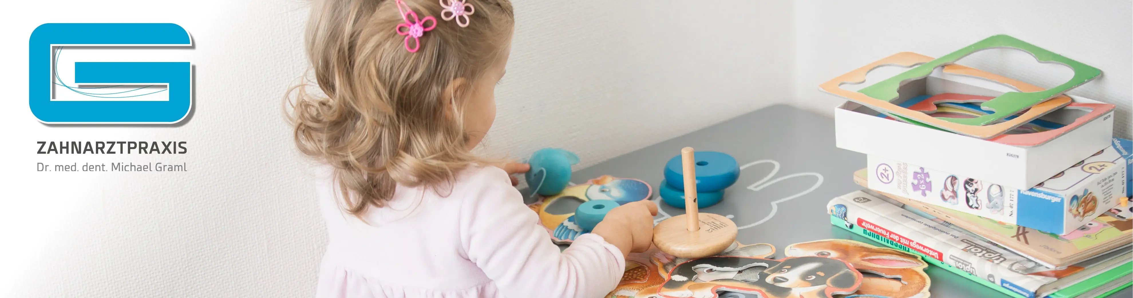 Das Bild zeigt eine freundliche und kindgerechte Szene in der Zahnarztpraxis Dr. med. dent. Michael Graml, wie links am Logo und dem Schriftzug zu erkennen ist. Im rechten Bildbereich spielt ein kleines Mädchen mit Spielzeug und Puzzles an einem Kindertisch. Sie trägt eine rosa Kleidung und hat zwei rosafarbene Haarspangen in Blumenform im Haar. Auf dem Tisch befinden sich verschiedene Spiel- und Lernmaterialien, darunter Holzspielzeug, Puzzleboxen und Kinderbücher. Die Szene vermittelt eine angenehme, familiäre Atmosphäre und zeigt, dass in der Praxis auch auf die Bedürfnisse kleiner Patienten eingegangen wird.